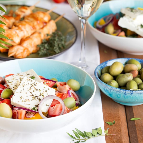 A Mediterranean spread with Greek salad, olives, chicken skewers, and white wine on a wooden table.
